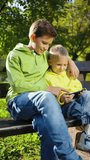 Vertical Screen: Two boys sitting on bench in park, watching funny video, playing online game, browsing internet on phone outdoors. Leisure time with gadget, addicted kids. Concept of technology