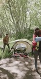 Vertical video of biracial couple pitching tent in forest, slow motion. Spending quality time, lifestyle, nature, hiking and camping concept.