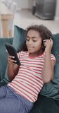 Vertical video of happy biracial girl using headphones and smartphone, slow motion. Spending quality time at home, domestic life and childhood.