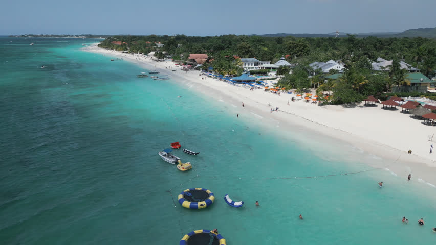 Aerial video of tourists on a beach with boats in the sea against sky, above people and water on a sunny day - Montego Bay, Jamaica