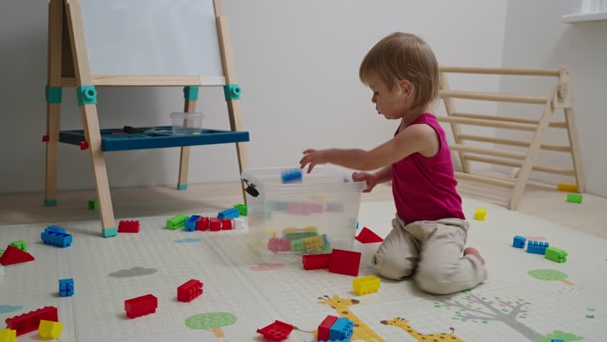 The child himself cleans up toys in the nursery after the game. Toddler baby collects toys in a box. Kid girl aged 2,5 year old.