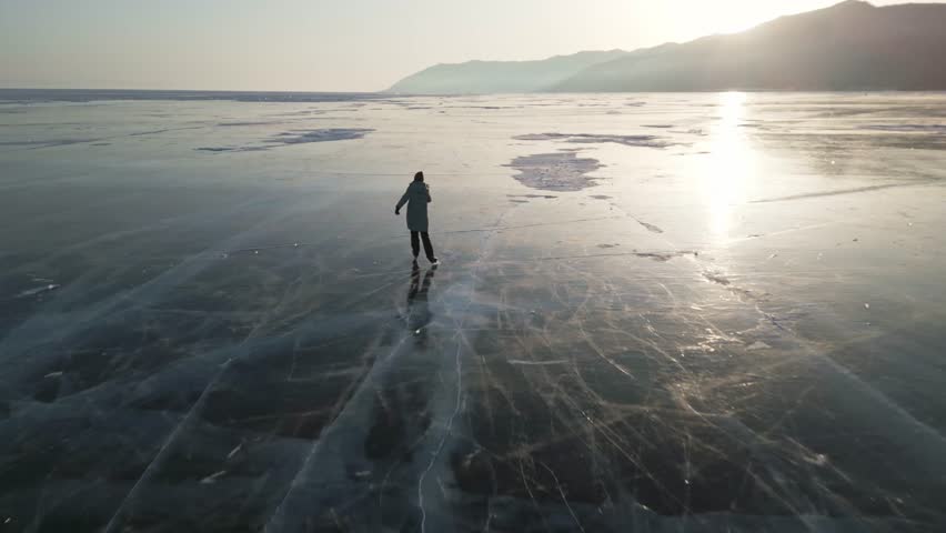 A woman in a blue down jacket skates on the blue transparent ice of the endless winter lake Baikal at sunset
