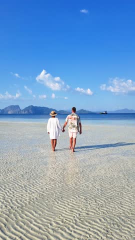 A couple holding hands strolls along the sandy beach, with the horizon stretching out over the calm waters. People relax under the cloudy sky