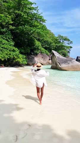 A woman wearing a white dress and hat strolls leisurely on the sandy beach, with the water glistening under the clear blue sky and lush trees in the background Similan Islands, Thailand Royalty-Free Stock Footage #3455964183
