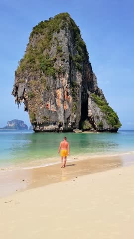 A man strolls along the sandy beach, near a towering rock. The vast body of water stretches to the horizon under the clear blue sky, surrounded by lush plants and trees Railay Beach, Krabi, Thailand Royalty-Free Stock Footage #3455970289