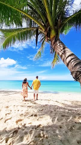 A man and a woman are standing on a sunny beach under a tall palm tree, surrounded by azure water and a clear blue sky Koh Kood, Thailand