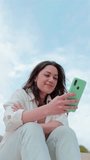 Vertical individual portrait of young adult brunette woman watching content on a cellphone app sitting outdoors. Carefree lady smiling and having fun texting messages with a smart phone. Copy scpace