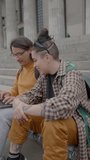 Young boyfriend and girlfriend having discussion and doing homework on laptop outdoors on staircase in front of the university. Vertical format clip