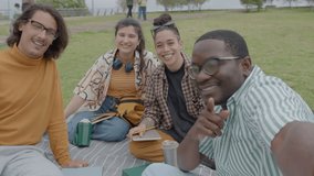 Group of cheerful college friends sitting on green lawn in the park, laughing and chatting on camera during online video call or live stream on social media