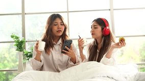 Two Asian female friends wearing white shirts sitting on the bed, waking up on a holiday morning, feeling lazy, drinking coffee and eating breakfast together in the apartment bedroom.