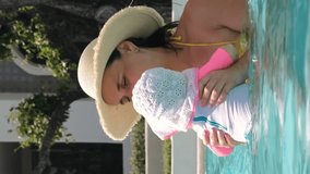 Vertical video. Young mother in straw hat resting with her baby in an outdoor pool durning vacation, slow motion