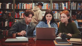 Three students engrossed in group study session in library with laptop and books on table amidst bookshelves - Powered by Shutterstock - Get 15% off with code: PIKWIZARD15