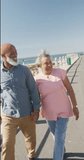 Vertical video of senior biracial couple walking along promenade by the sea, slow motion. Spending quality time, lifestyle, vacation and retirement concept.