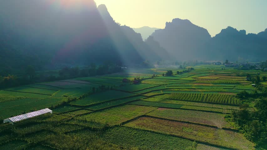 Mystic fertile valley and patchwork crops fly over reflective pond, with soft evening sun rays in mountainous horizon. Vang Vien, Laos.