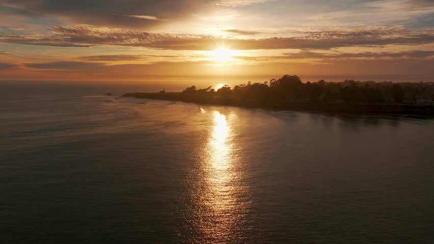 Drone shot flying in towards a light house at sunset with hundreds of surfers