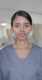 Vertical video of portrait of happy biracial female nurse looking at camera, in slow motion. Hospital, medicine and healthcare.