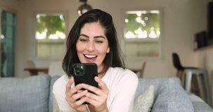 Beautiful young Mexican woman sit on couch with modern smart phone, laughing, enjoy pleasant chat, typing answer, joking, having fun while communicate in social media with friend on weekend at home