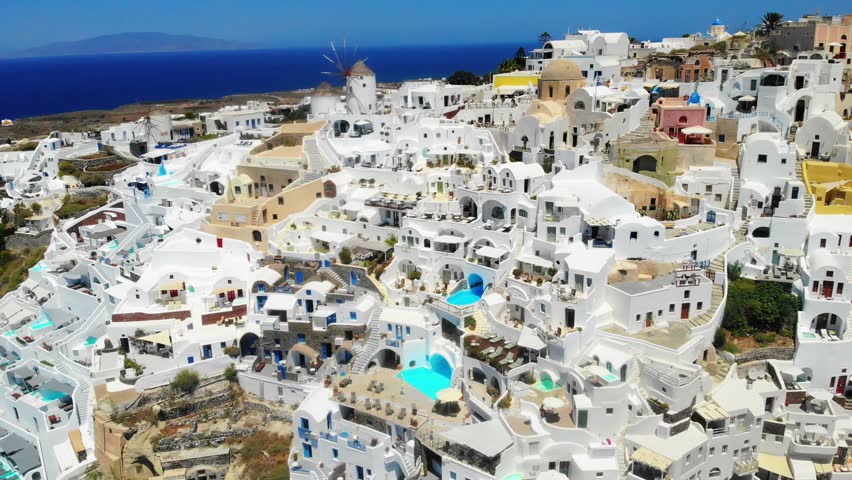 Breathtaking aerial footage of a charming coastal town on a hill with white buildings, a windmill, and a stunning blue ocean backdrop in Santorini, Greece
