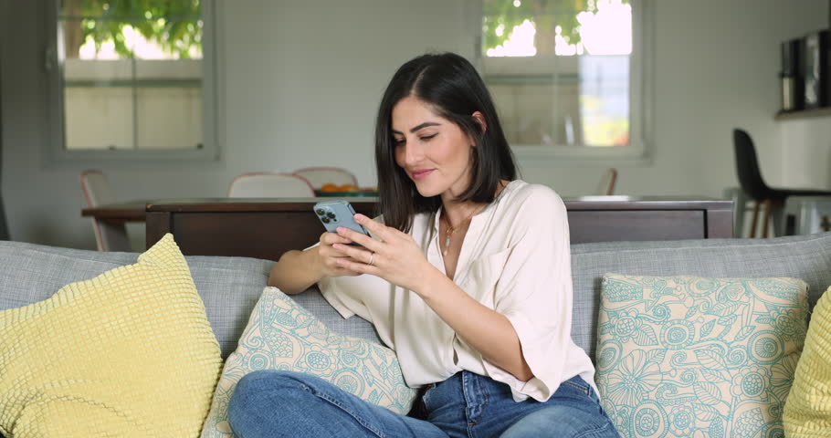 Attractive Latina woman sit on sofa with smartphone, laughing, enjoy on-line chat, spend weekend share messages with friend, enjoy e-dating services for singles, Modern tech, remote communication, fun Royalty-Free Stock Footage #3456292511