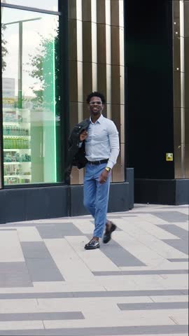 Fashionable African American man walks in city, wears stylish outfit blue trousers and shirt, loafers and leather jacket. Street style male fashion