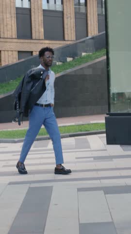 Full length Fashionable African American man walks in city, wears stylish clothes blue trousers and shirt, loafers and leather jacket. Street style male urban fashion