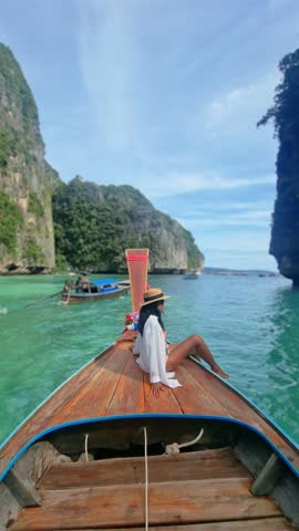 A woman is perched on the bow of a boat, gliding through the tranquil ocean water under a cloudy sky. The natural landscape is serene as she travels on the watercraft to Koh Phi Phi Thailand