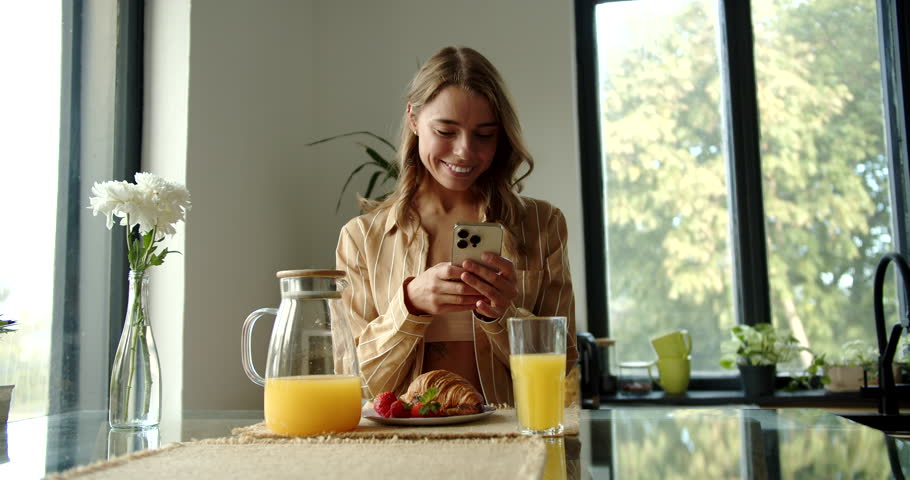 woman sitting at the table in the kitchen, smiling and looking at the phone