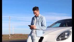 Video of curly haired Caucasian man leaning on his car smiling, using mobile phone, with the road beside, side view. Lifestyle concept