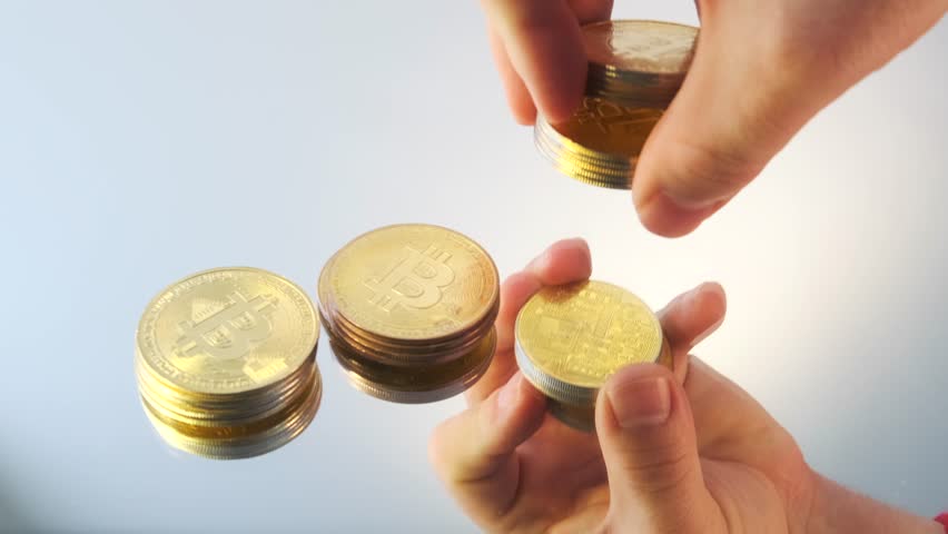 Gold bitcoin coins in man's hand divides into three parts on background of mirror surface white table, close up. Bitcoin cryptocurrency. 