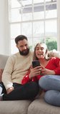 Vertical video of happy caucasian couple sitting on sofa and using smartphone. Lifestyle, love, relationship and spending time together concept.
