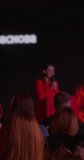 A woman in a red suit performs in a dark room in front of a group of people. A stand-up performance by a millennial woman, an unrecognizable face. Vertical video. High quality 4k footage