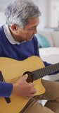 Vertical video of senior biracial man singing and playing acoustic guitar, sitting at home. Retirement, hobbies, domestic life, inclusivity and senior lifestyle concept.