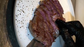 a cook with metal tongs places sliced grilled beef steak on a plate and adds corn and red chili peppers to it. Juicy sliced ribeye steak with salt. Delicious meat food concept. vertical video