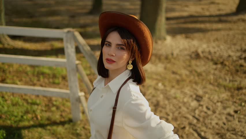 Sunset Ranch Elegance. Woman in hat gazes into the distance at a ranch during golden hour.