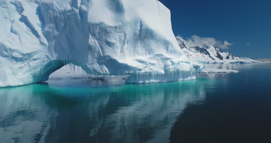 Melting arch in big glacier in Antarctica. Massive iceberg with icicles reflected in calm water. Ecology, melting ice, climate change, global warming. Low angle drone flight over crystal ocean closeup