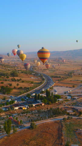 Vertical video. In this aerial video, the skies above Cappadocia, Turkey, come alive with a kaleidoscope of hot air balloons. Against the backdrop of the region's iconic valleys, rocks, and fields