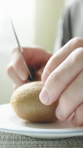 A man cuts and eats a kiwi fruit with a spoon, in the sun, close-up. Vertical video.