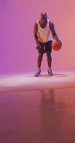 Vertical video of african american male basketball player bouncing ball on pink background. Sports and competition concept.