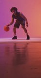 Vertical video of biracial male basketball player with ball on pink background. Sports and competition concept.