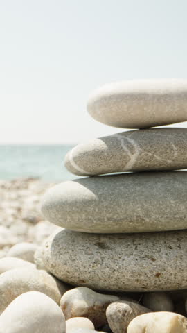 Close-up, a woman makes a cairn. Handmade stone tower on the seashore. A stack of balanced pebbles on the beach. Against the backdrop of the sea and people swimming Vertical video.