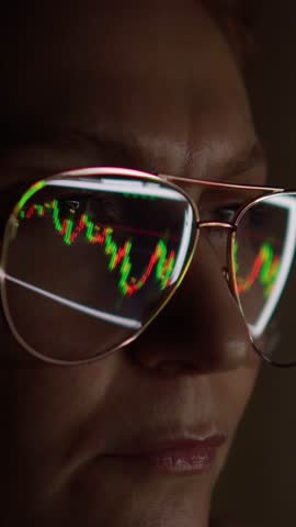 A trader girl trades forex at night at home. The computer screen with stock charts and market analysis is reflected in the glasses on his face. Vertical video for shorts. Royalty-Free Stock Footage #3456928519