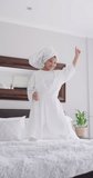 Vertical video of happy biracial girl in robe jumping on bed. Childhood, fun and spending time at home concept digitally generated video.