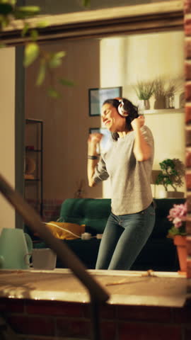 Vertical Screen: Happy Hispanic Woman Wearing Wireless Headphones Dances In Her Cozy Apartment. Cheerful Lively Girl Listening to Music at Home and Dancing in a Good Mood. Shot Inside Apartment Window