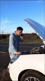 Video of irritated curly haired Caucasian man talking on the phone worried looking at the engine of his broken car, vertical video. Road emergency concept