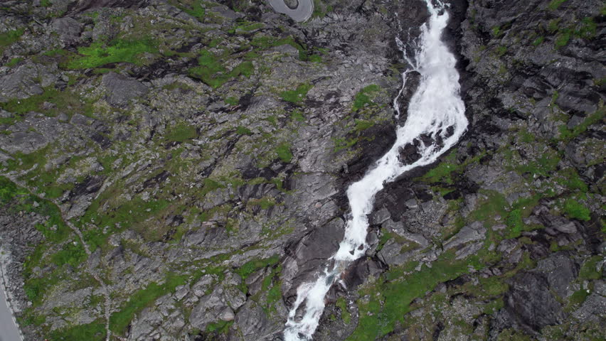 Top down aerial shot of the Trollfossen waterfall cascading down a cliff into the Tverelva river in Norway. Hairpin bends of the serpentine Trollstigen mountain road can be seen next to the waterfall. Royalty-Free Stock Footage #3457105629