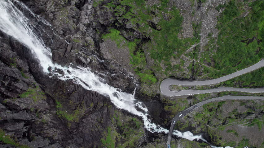 Bird's eye Aerial view showing the tight hairpin bends of the Trollstigen road in Norway. The Stigsfossen waterfall can be seen. Many cars drive on the road. Loose rocks and trees by the road. Royalty-Free Stock Footage #3457111557