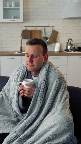 Folk medicine. Sick young man with fever wrapped in warm blanket drinking hot tea. Pandemic. Vertical video.