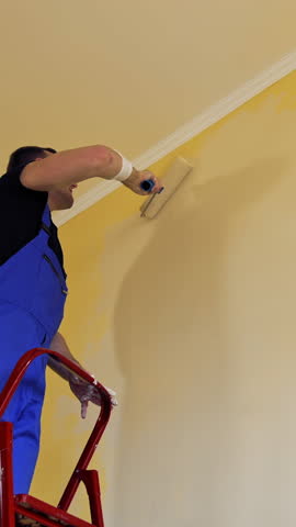 Man painting a yellow wall with a roller at work. Offering professional painting services. Vertical video.