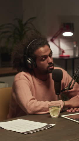 Vertical of Biracial guy visiting dark broadcast studio at nighttime, giving interview during audio podcast recording