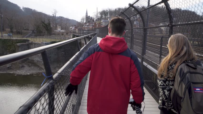 Camera follows teenage son and blonde mature woman walking across a foot bridge over the Shenandoah River towards Harpers Ferry National Park.
